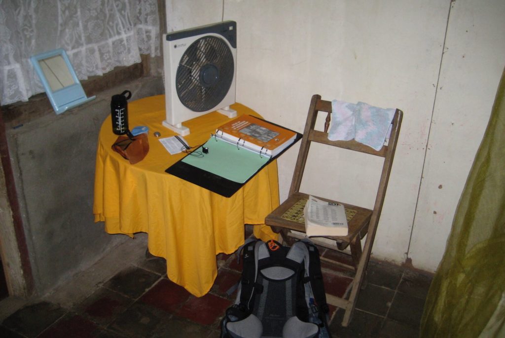 The view of my desk in Nicaragua as a Peace Corps volunteer. You can see concrete stone block walls, a small table with a cover over it, a fan, a foldout chair, and the edges of a mosquito net. A simple existence. Lifestyle creep started from a low level for me!